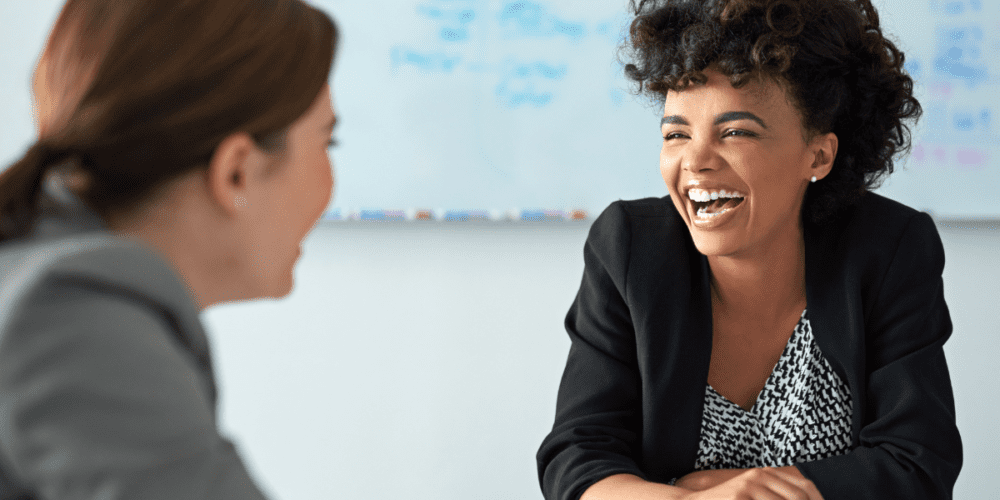 Two women coworkers talking at a table and laughing.